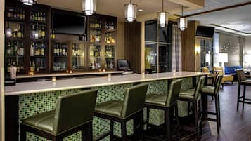 A bar area with a long counter and multiple stools, a television screen, and a variety of bottles and glasses on shelves.