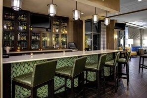A bar area with a long counter and multiple stools, a television screen, and a variety of bottles and glasses on shelves. - Holiday Inn Indianapolis Carmel by IHG (Indianapolis)