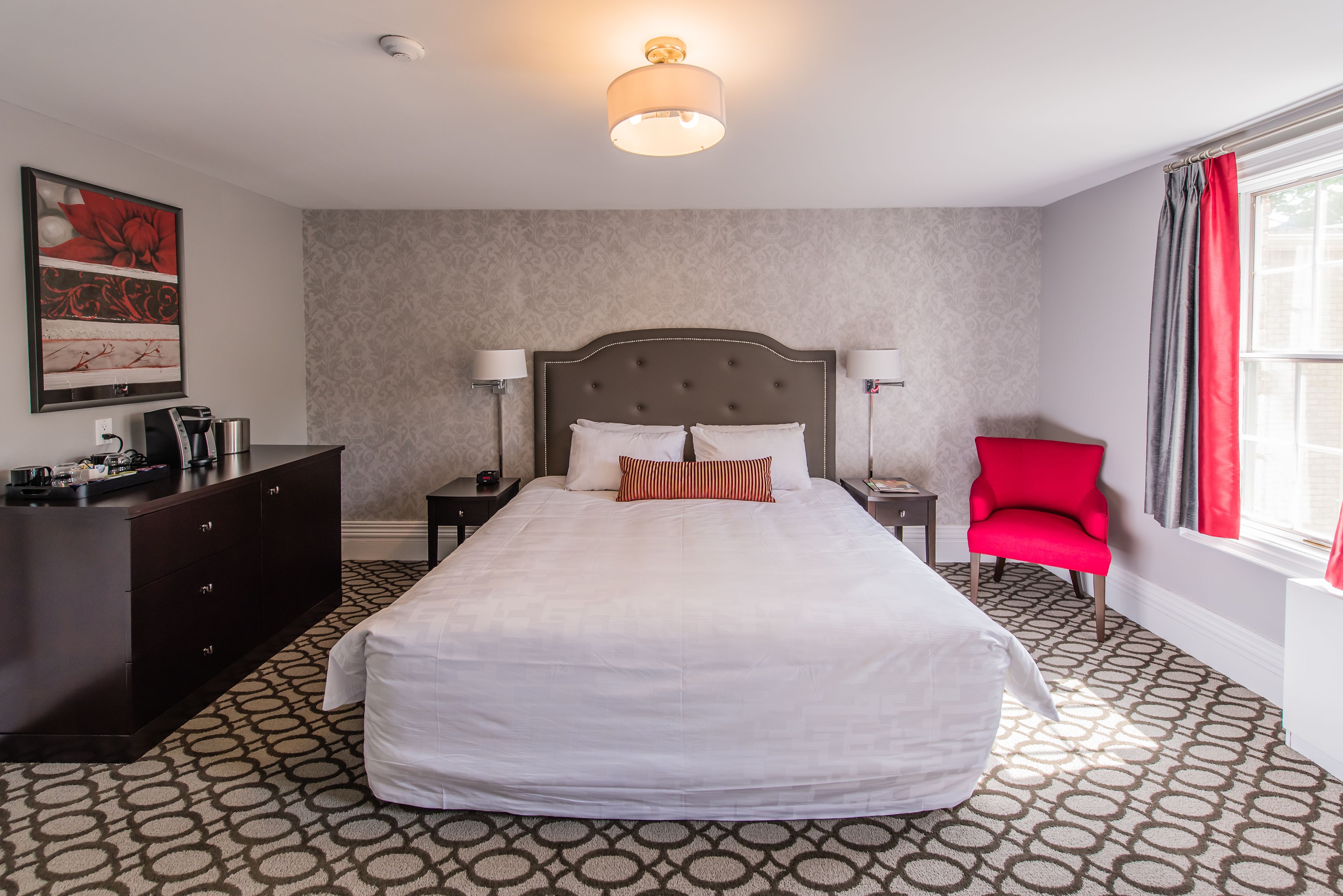 Une chambre d’hôtel avec un grand lit, une commode, deux tables de chevet, un fauteuil rouge et une fenêtre avec des rideaux.