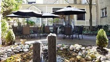 Une terrasse extérieure avec des sièges en pierre et des parasols, une petite fontaine et un bâtiment en arrière-plan.