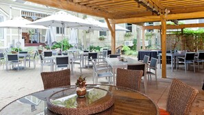 Un patio al aire libre con mesas redondas de vidrio y sillas de mimbre, rodeado de sombrillas y una pérgola de madera.