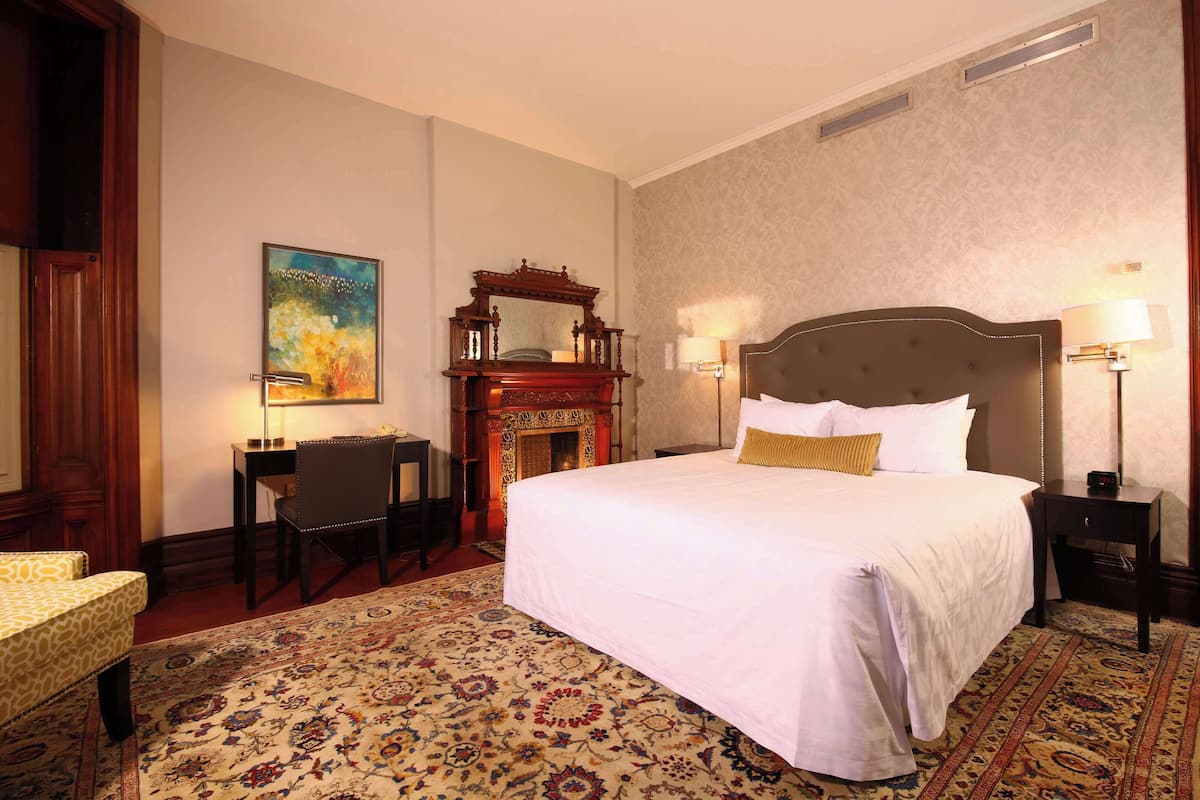 A hotel room with a large bed, a desk with a chair, a fireplace, and a painting on the wall.