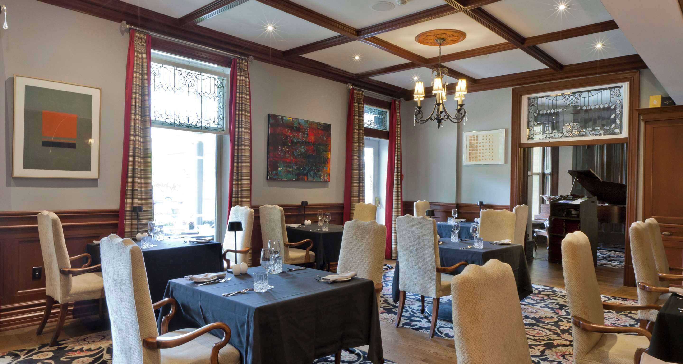 A dining area with tables set for four, wooden chairs, a chandelier, and artwork on the walls.