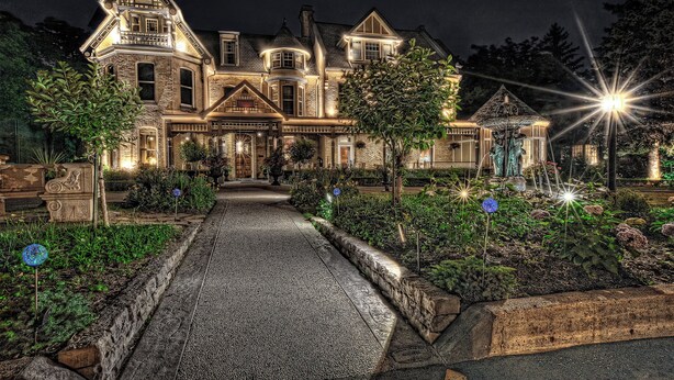 Una casa grande de estilo victoriano, iluminada, con una entrada principal destacada, un jardín bien cuidado y un camino de entrada pavimentado.