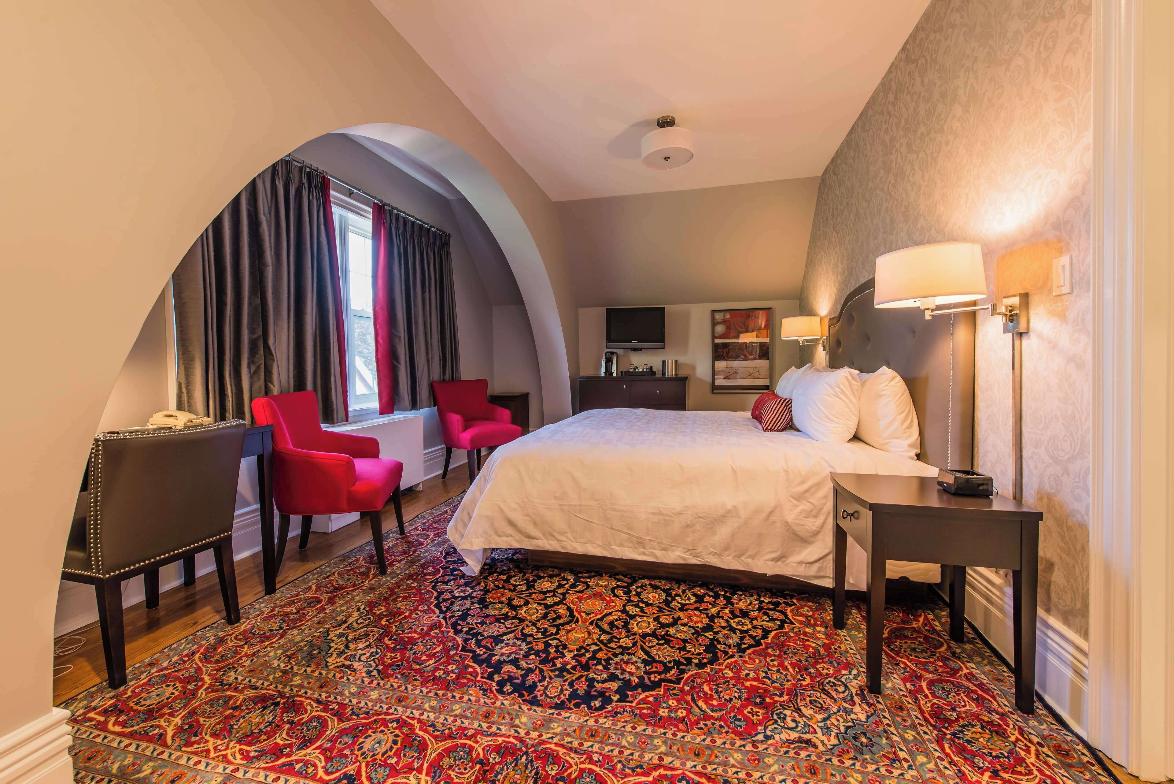 A hotel room with a large bed, a red armchair, a small table, a TV, and a patterned carpet.