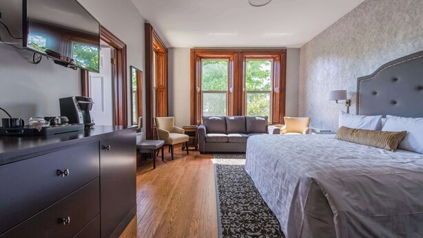 A modern hotel room with a large bed, a sofa, a desk with a coffee maker, and a view of trees through the windows.