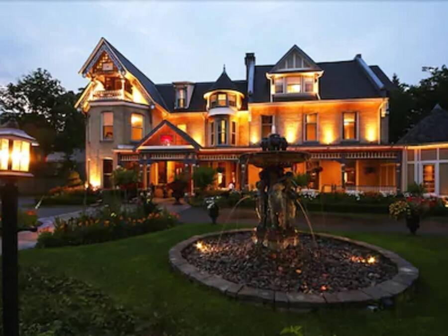 Uma casa grande, com vários andares, varanda frontal imponente e uma fonte no jardim.