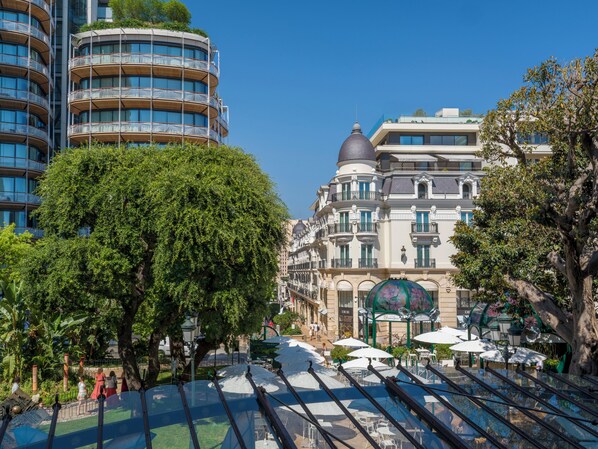 A modern building with multiple balconies, a large tree in the foreground, and outdoor seating with umbrellas.