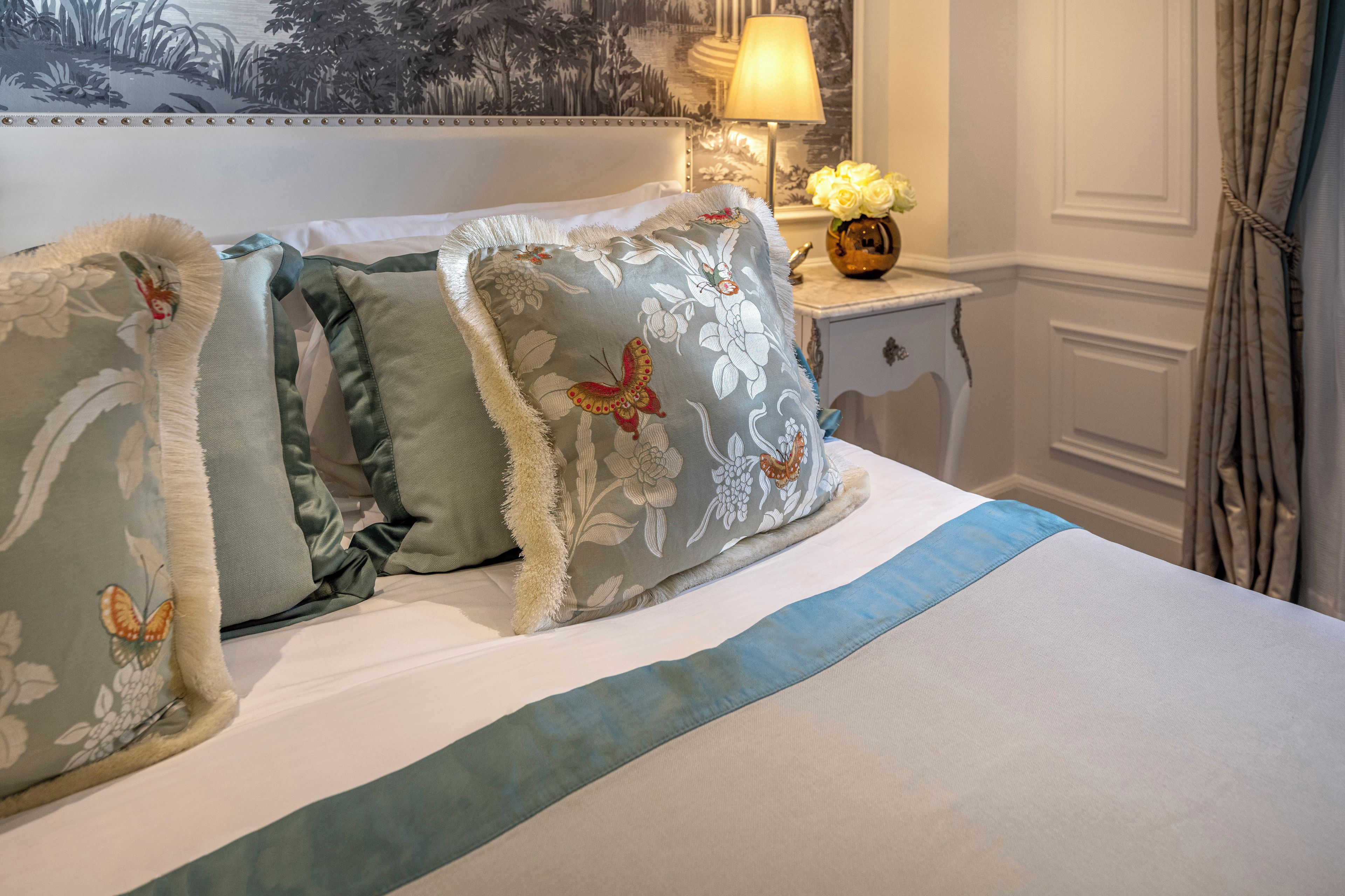 A neatly made bed with floral and butterfly-patterned pillows, a bedside table with a lamp and vase, and a wall with a nature-themed painting.
