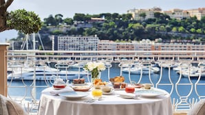 Mesa dispuesta para el desayuno con croissants y fruta, con vista a un puerto deportivo y edificios al fondo.