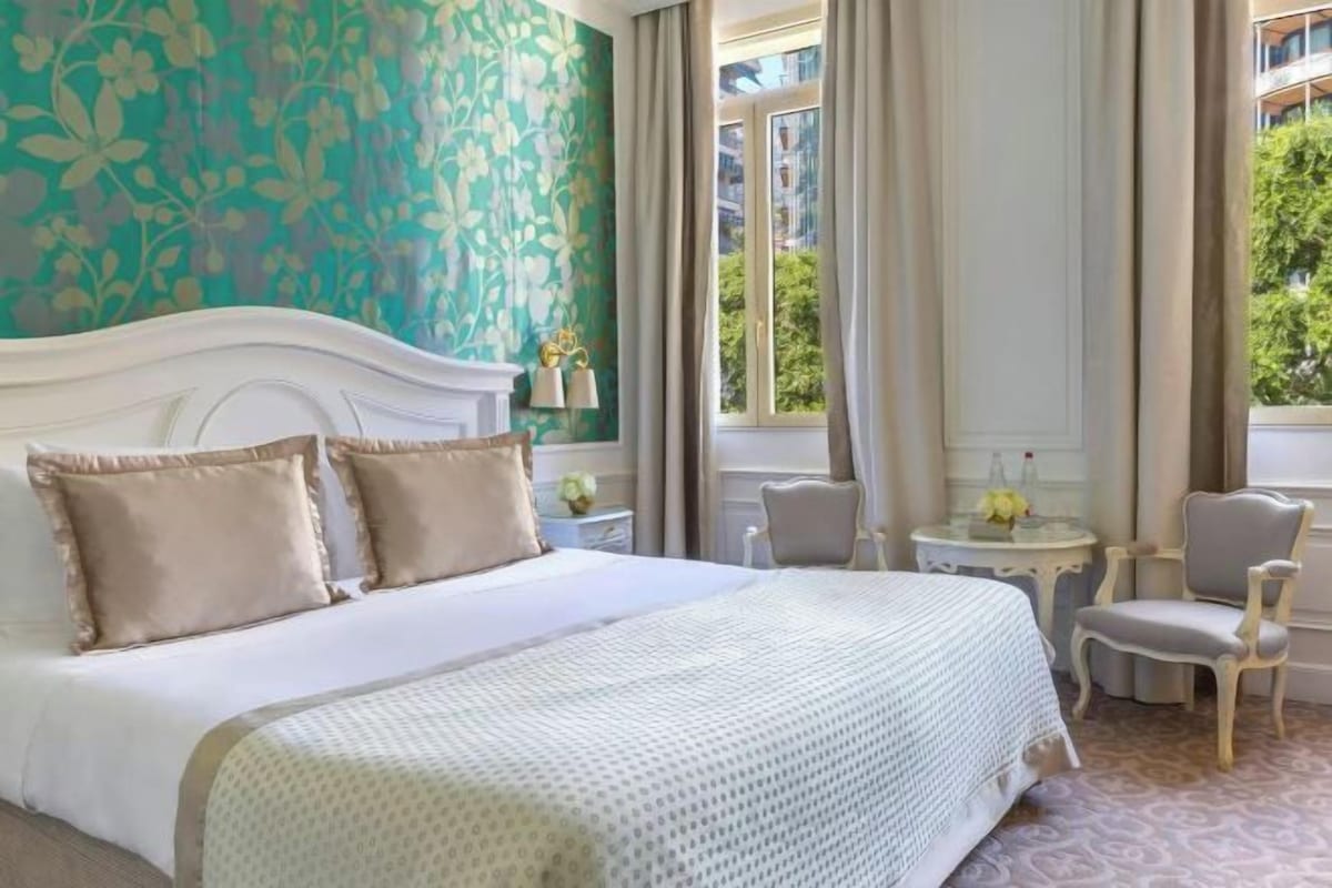 A hotel room with a large bed, two chairs, a small table, and a window with curtains.