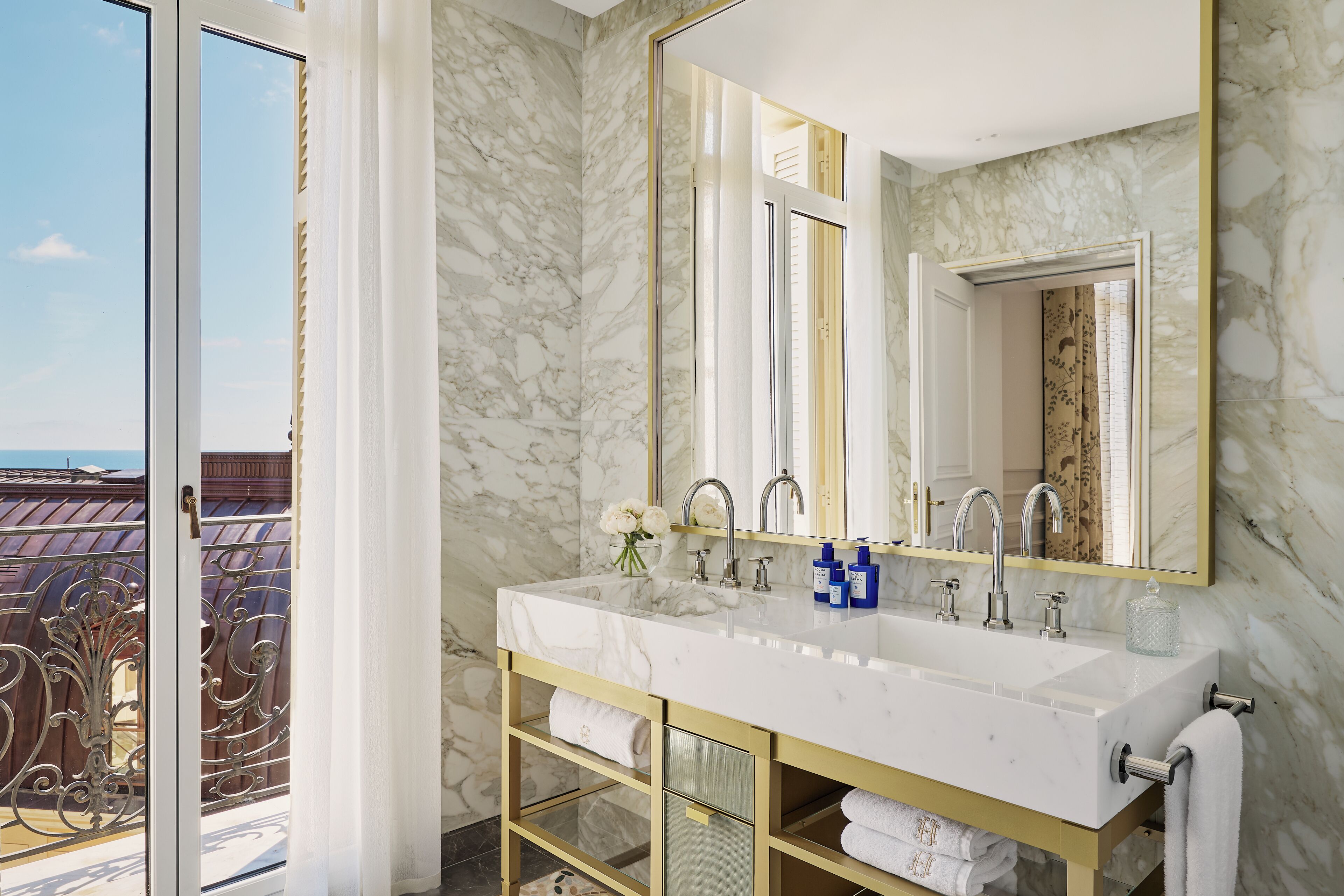 Un baño con pared de mármol, un espejo grande, un lavamanos blanco con dos grifos y un perchero para toallas.