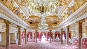 Un salón imponente con detalles ornamentales dorados, grandes candelabros y cortinas rojas.