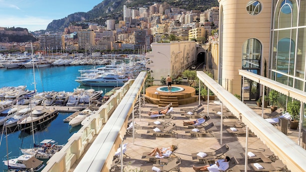 Terraza con sillones de sol y piscina, con vistas a un puerto deportivo con barcos y un paisaje urbano con una montaña al fondo.