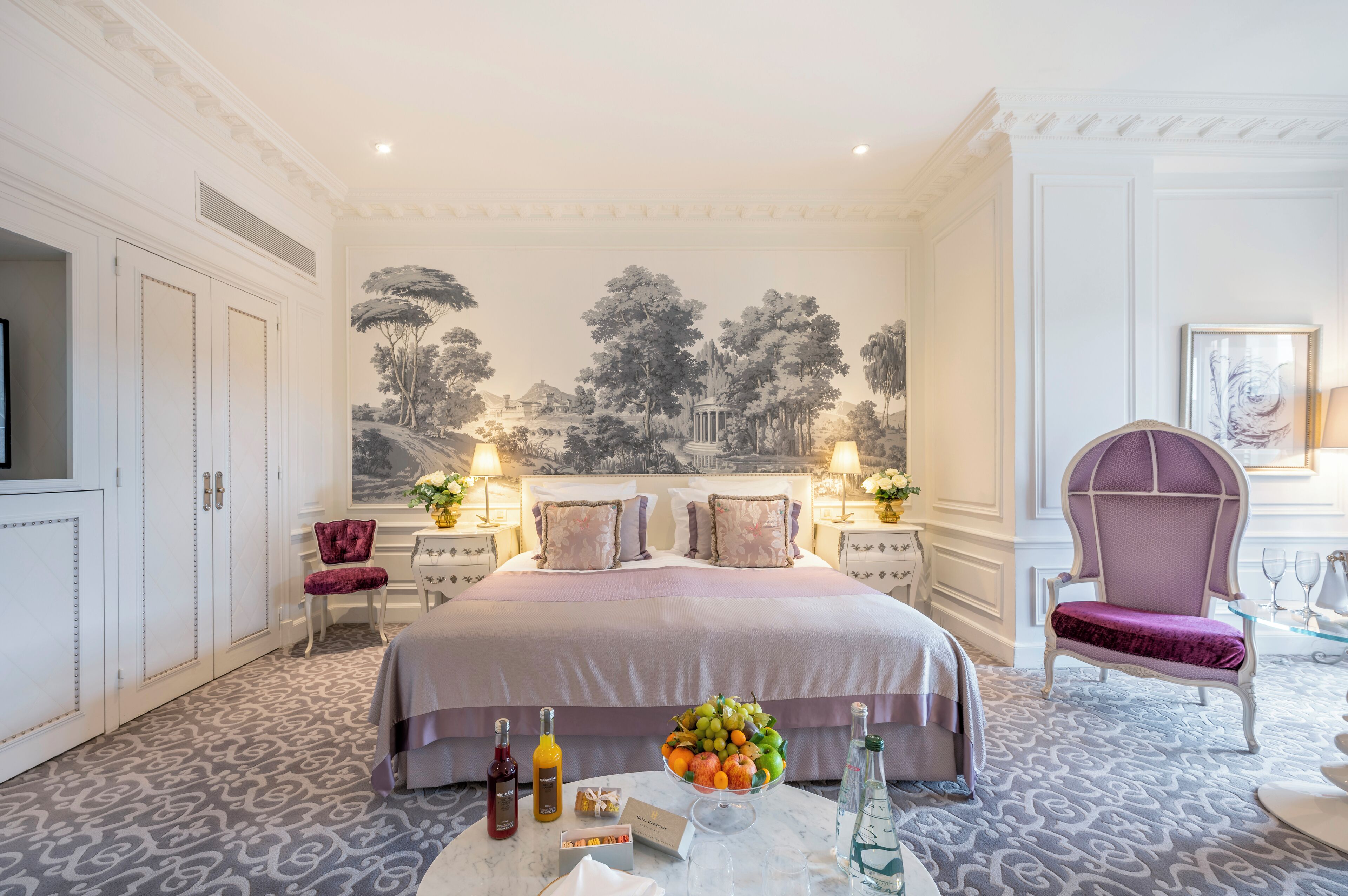 A hotel room with a large bed, a fruit bowl, and a table with bottles and glasses.
