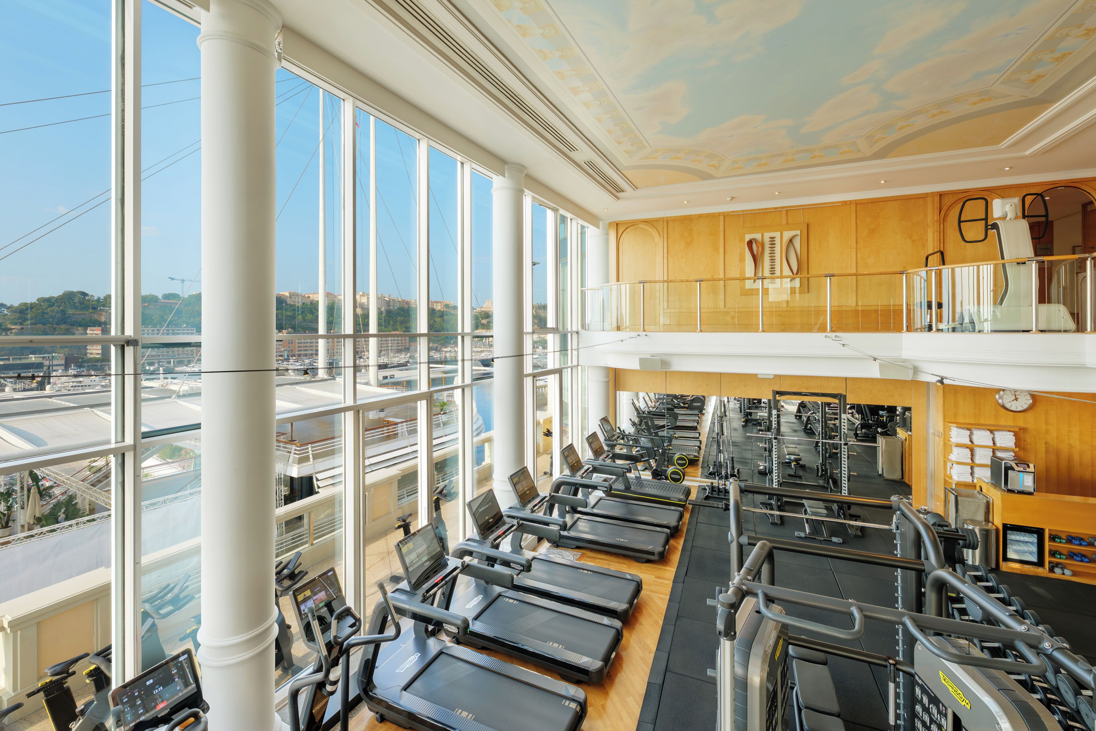 A gym with multiple treadmills, weight machines, and a view of the city through large windows.