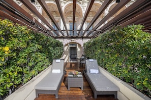 A sunlit outdoor lounge area with two reclining chairs and a small table, surrounded by lush greenery and a view of ornate architecture.