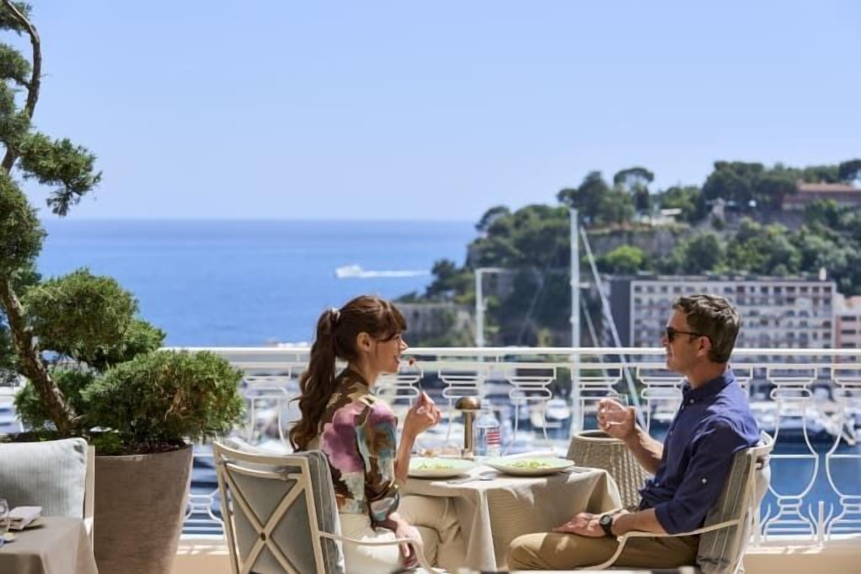 A couple dining at an outdoor table with a view of the sea and buildings in the background.