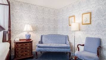 A bedroom with a four-poster bed, a wooden nightstand with a lamp, a blue patterned armchair, and a matching chair.