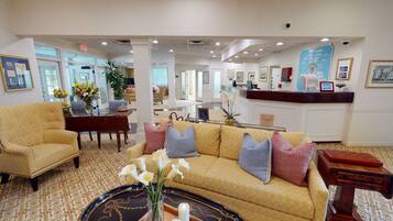 A hotel lobby with a reception desk, seating area with a patterned sofa, a coffee machine, and framed pictures on the walls.