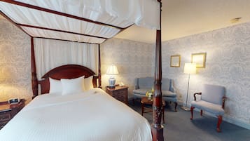 A four-poster canopy bed with a wooden headboard, a bedside table with a lamp, a chair, and a nightstand with a phone.