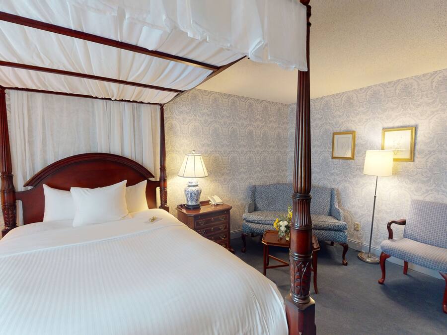 A four-poster canopy bed with a wooden headboard, a bedside table with a lamp, a chair, and a nightstand with a phone.