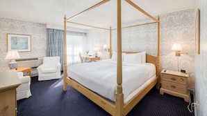 Un lit à baldaquin dans une chambre d’hôtel, avec une moquette bleue, un bureau avec une lampe, une chaise et une table de chevet avec une lampe.