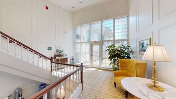 A spacious lobby with a high ceiling, a staircase, a patterned carpet, a yellow armchair, a marble table, and large windows offering a view of the outside.