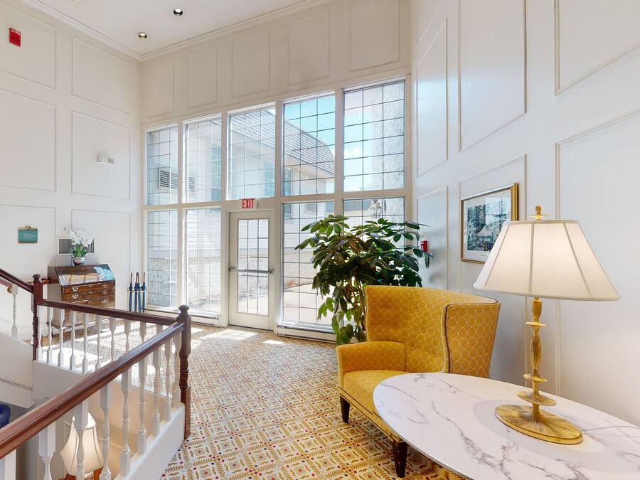 A spacious lobby with a high ceiling, a staircase, a patterned carpet, a yellow armchair, a marble table, and large windows offering a view of the outside.