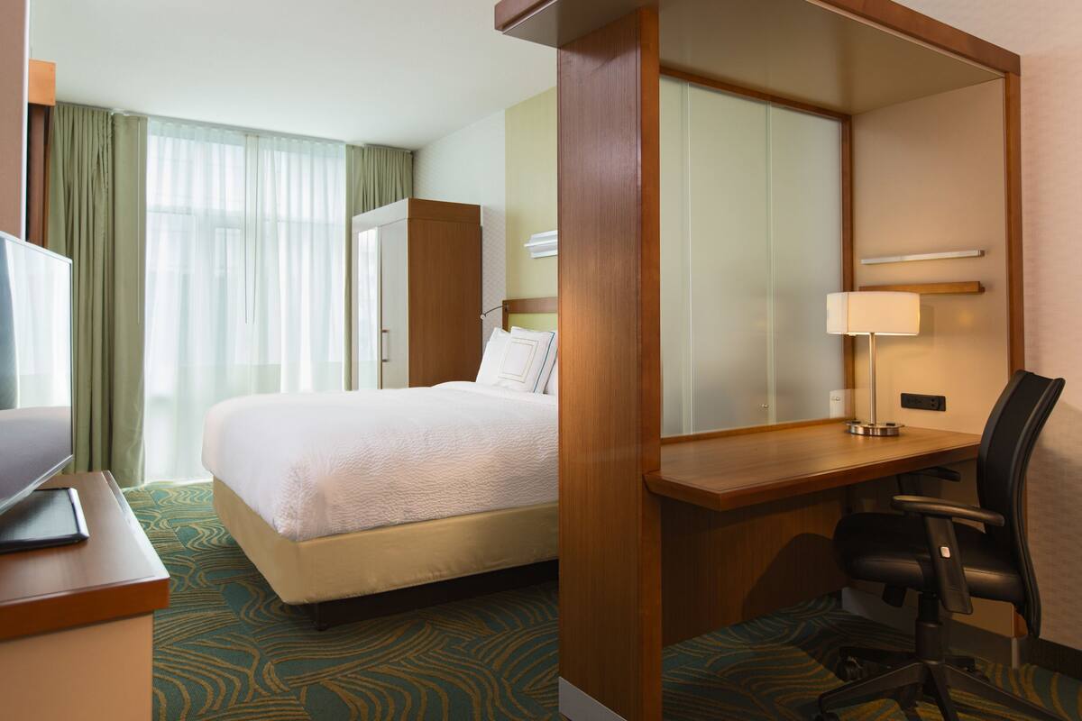 Ein Hotelzimmer mit Bett, Schreibtisch, Stuhl, Fernseher und einem Fenster mit Vorhängen.