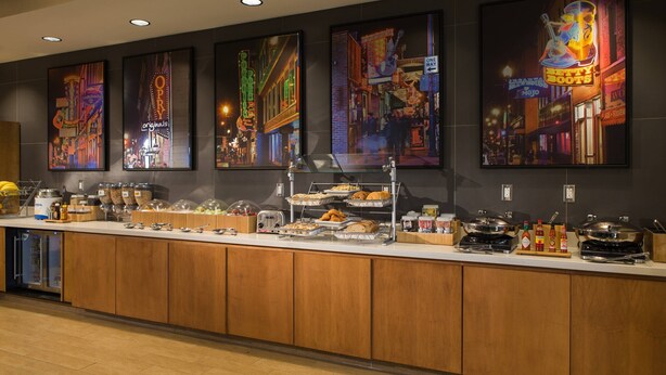 Un mostrador de buffet con diversos alimentos y condimentos, rodeado de obras de arte en la pared que representan escenas urbanas.