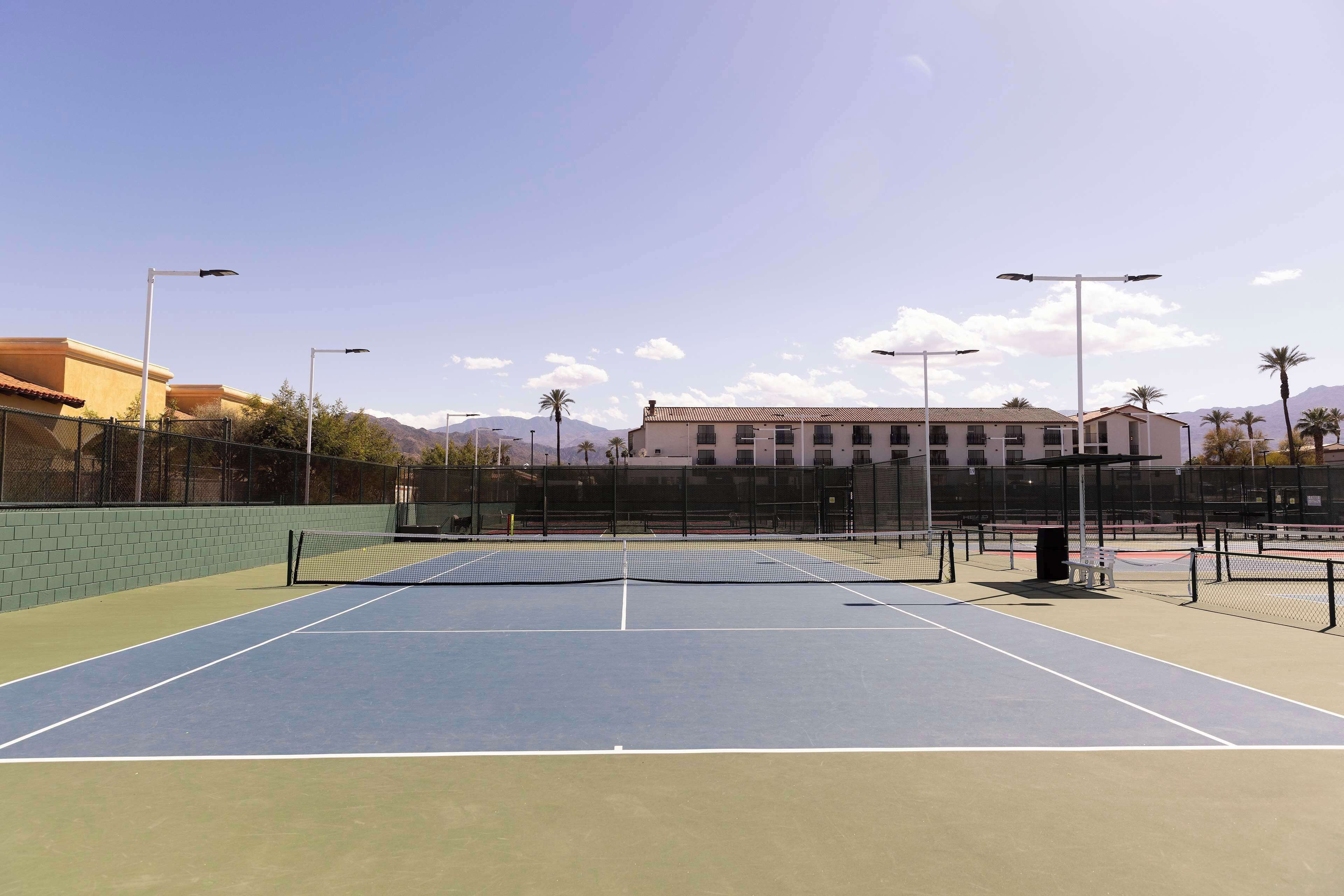 An outdoor tennis court with a net, surrounded by a fence, and a building in the background.