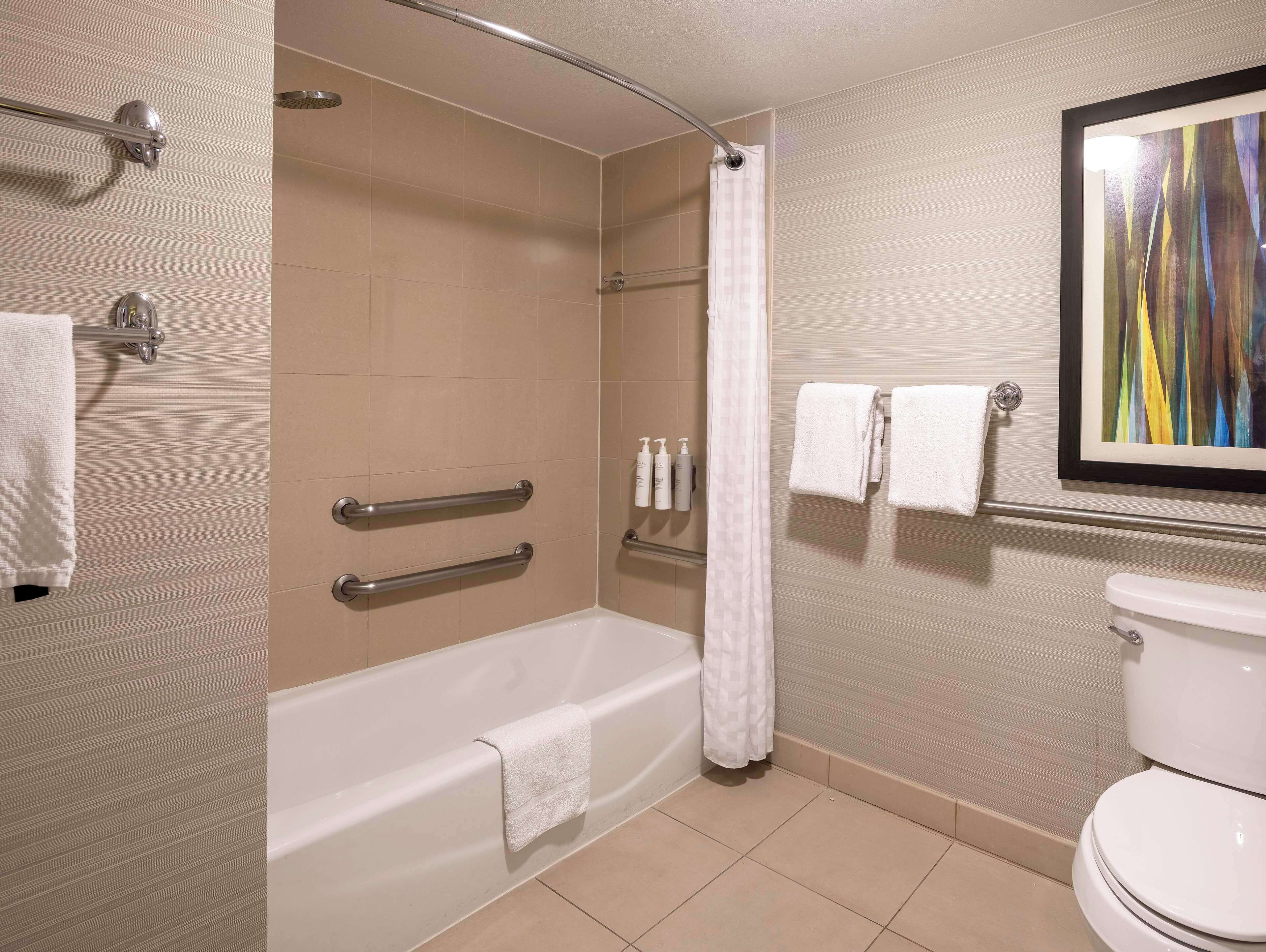 A bathroom with a bathtub, a shower, and a toilet. There are grab bars installed on the bathtub, a shower curtain, and a towel rack with towels.