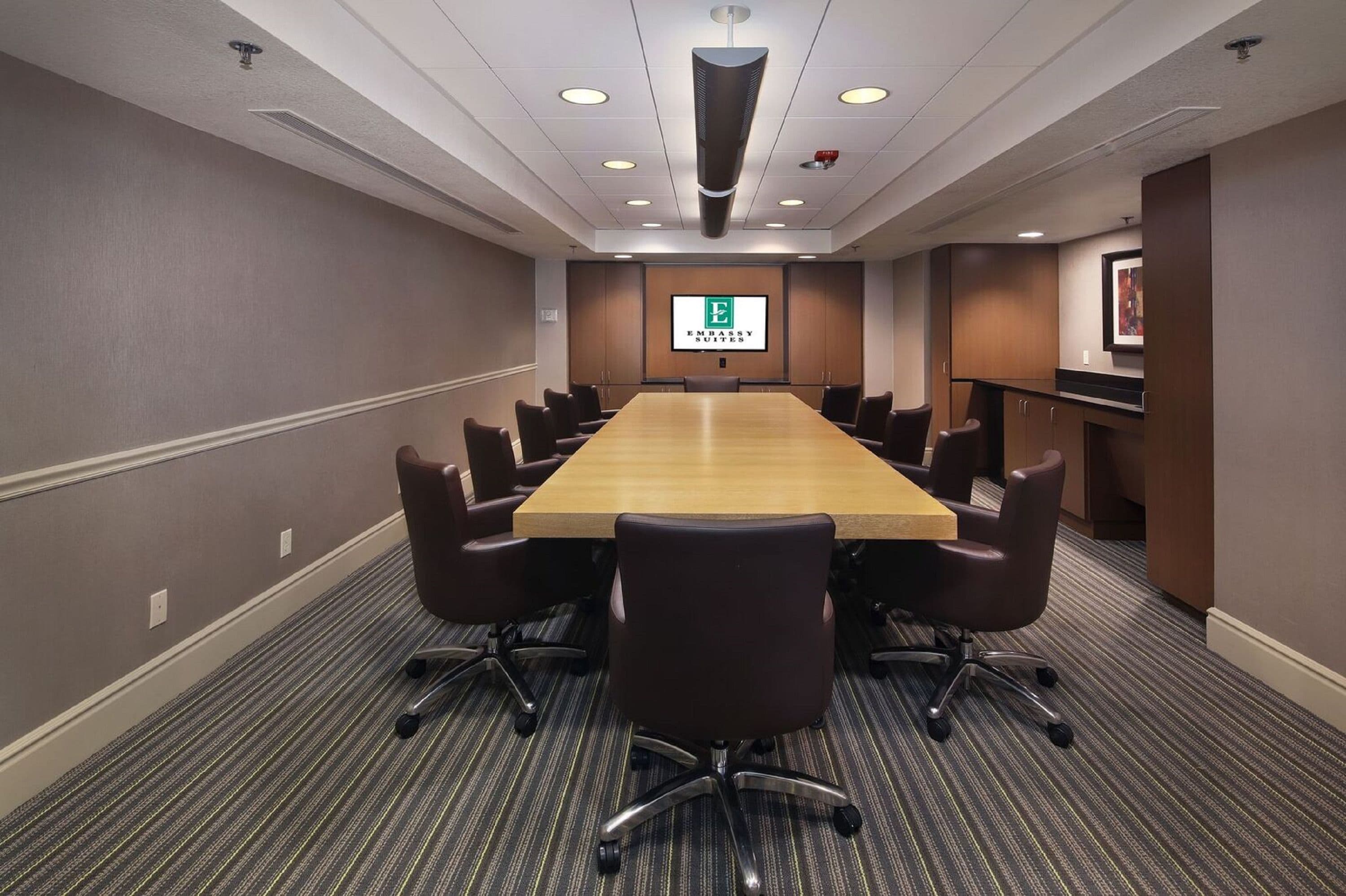 A conference room with a long table, multiple chairs, a projector screen, and a carpeted floor.