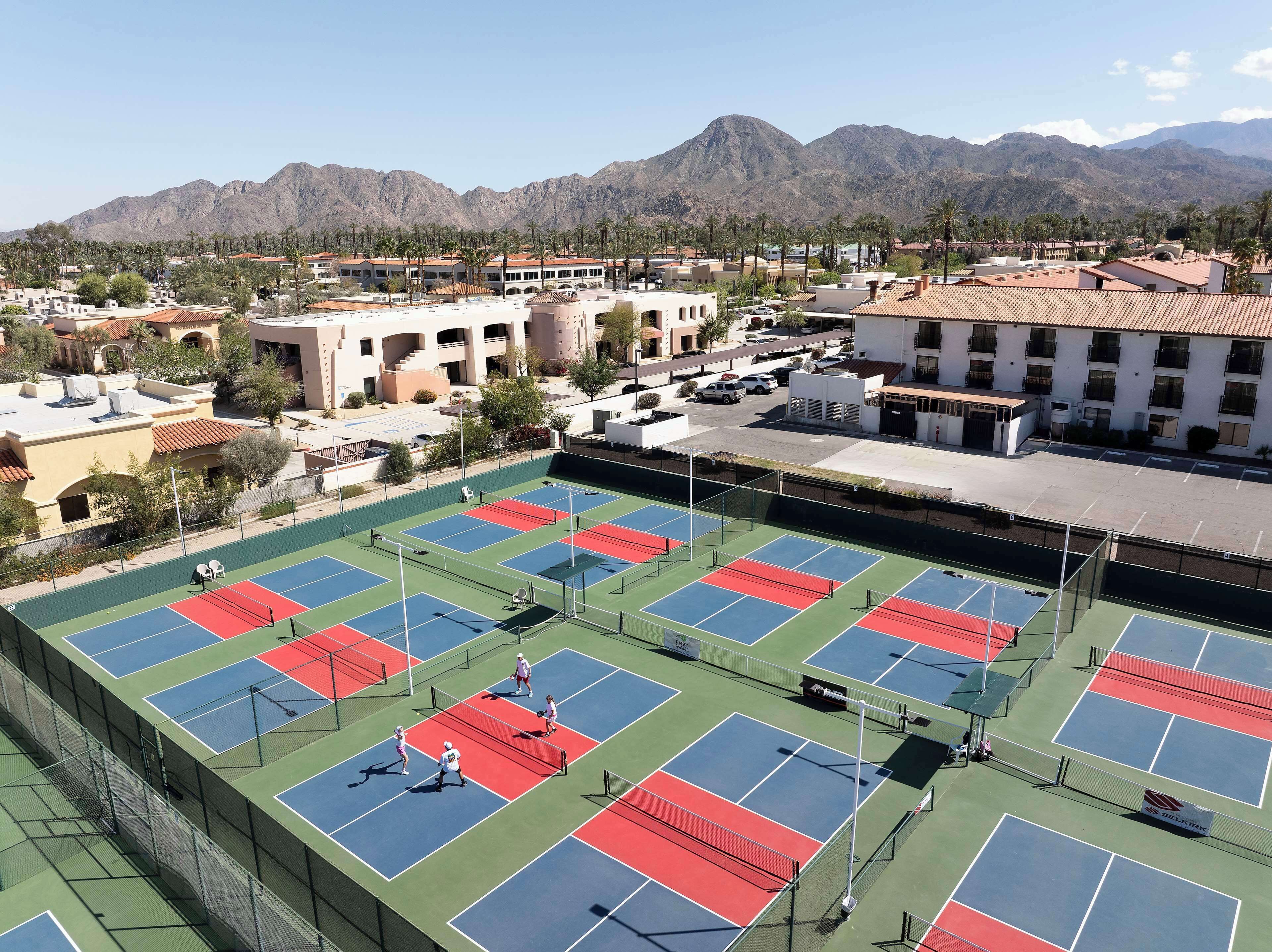 Complejo de canchas de tenis con varias pistas, rodeado de edificios y palmeras, con montañas al fondo.