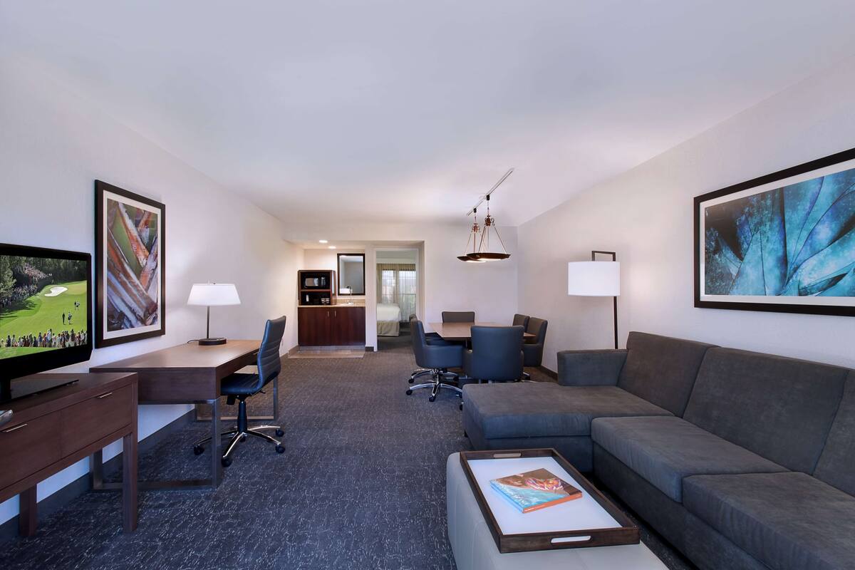 A hotel room with a TV, desk, office chair, sofa, dining table, chairs, and a kitchen area.