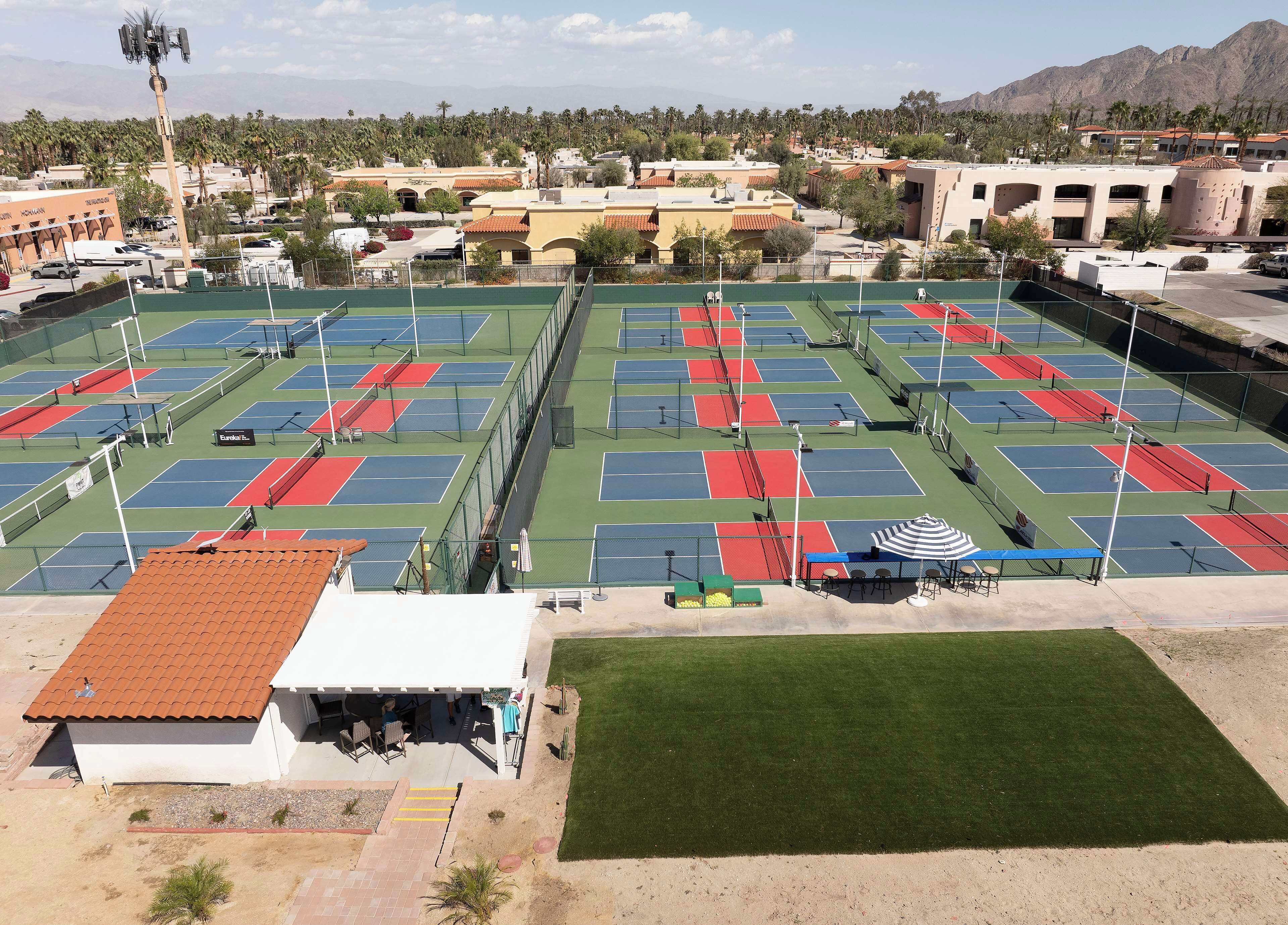 Vue aérienne d’un complexe de courts de tennis comprenant plusieurs terrains, un espace de sièges couvert et un bâtiment surmonté d’un toit en tuiles rouges.