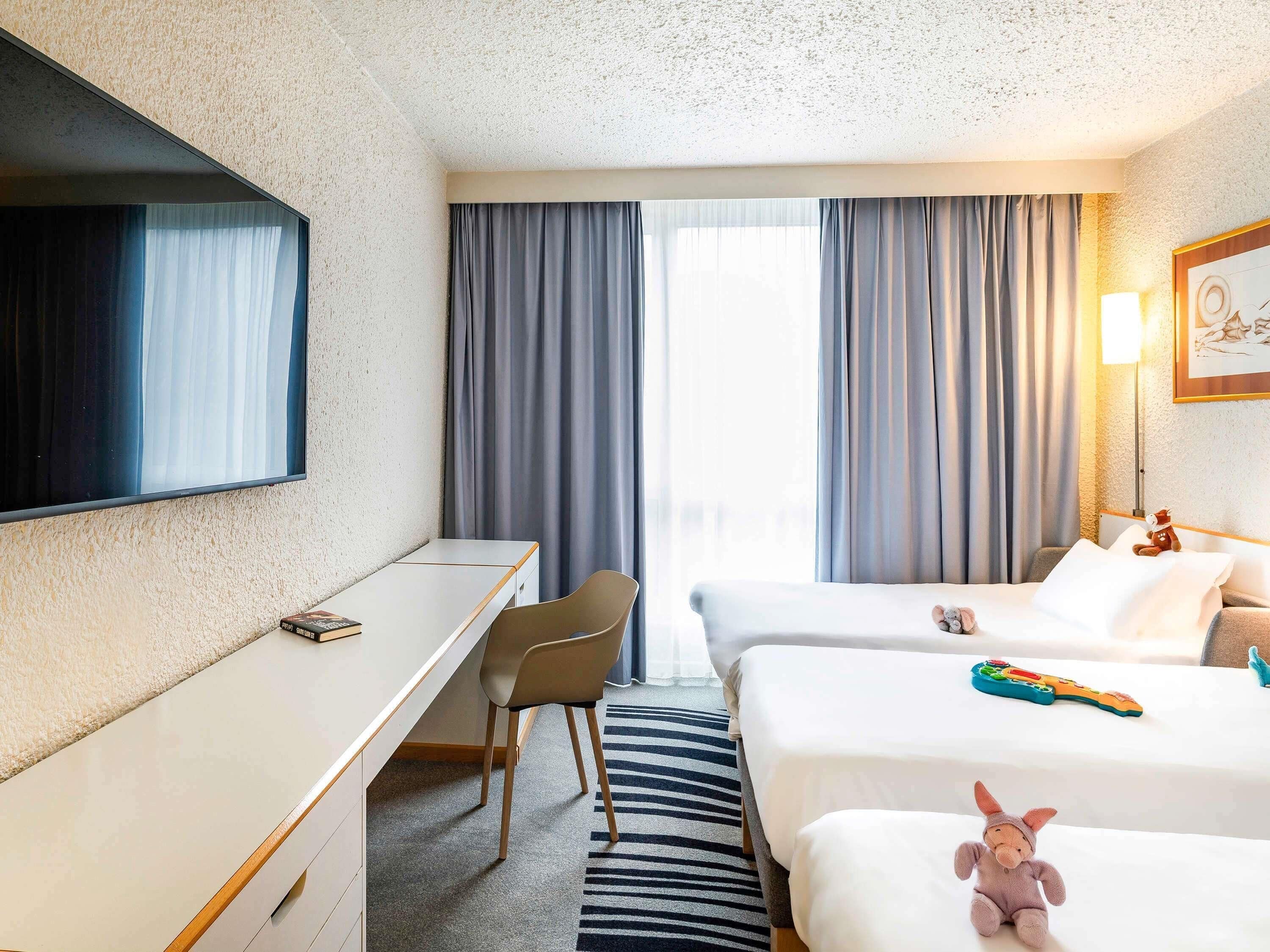 Et hotelværelse med skrivebord, to senge, et fjernsyn, en lampe og en bamse på sengen.