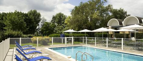 Una piscina con sillas reclinables azules y sombrillas, rodeada por una valla blanca y árboles.
