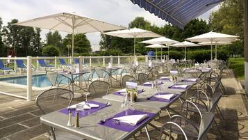 An outdoor dining area with tables and chairs, umbrellas, and a pool in the background.
