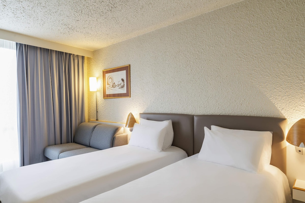 Une chambre d’hôtel avec deux lits, un bureau, une chaise, une lampe et un tableau encadré au mur.
