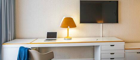 A hotel room with a white desk, a laptop, a lamp, a TV, a chair, and a bed.