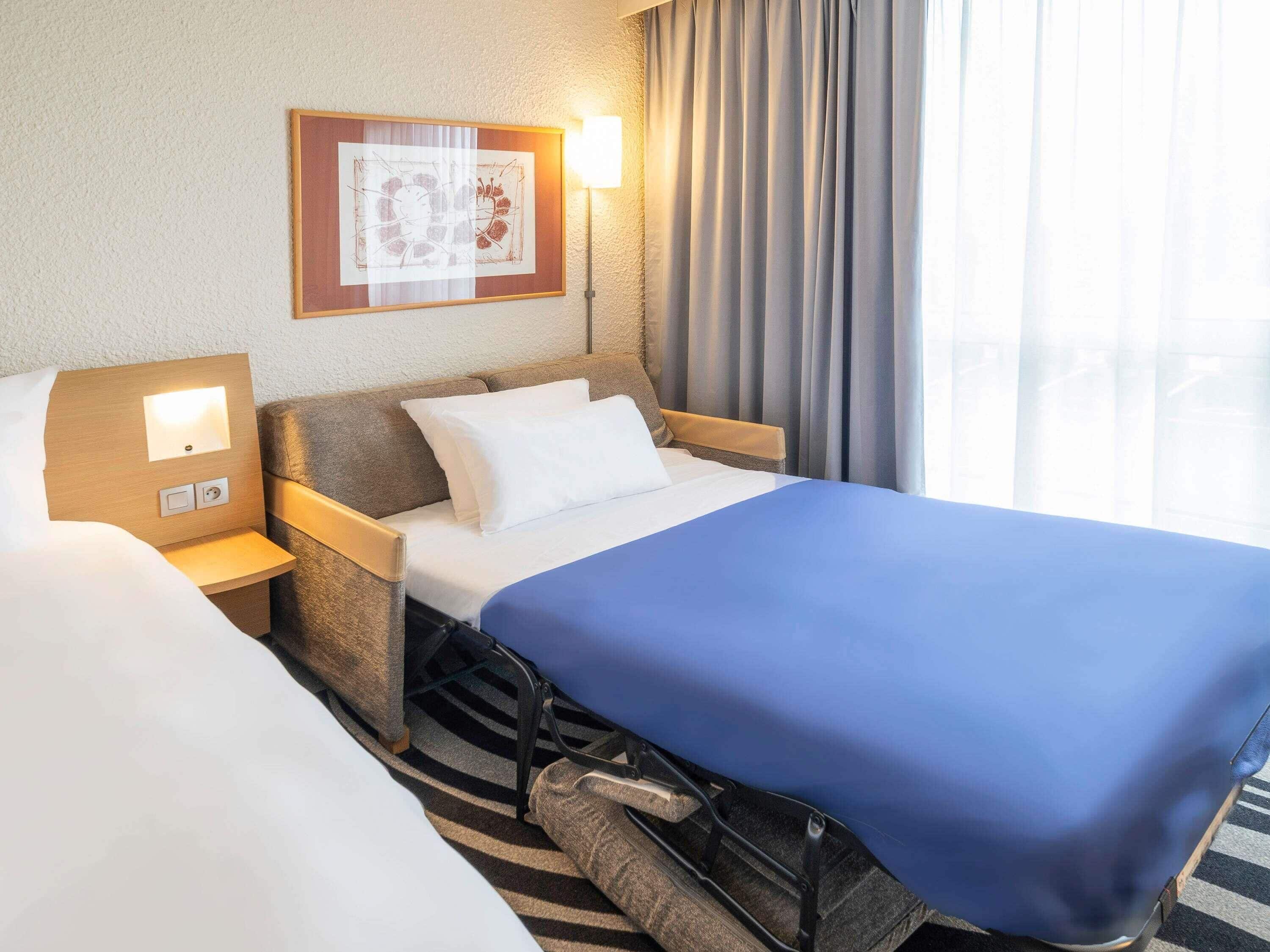 A hotel room with a bed, a nightstand, a lamp, a framed picture, and a window with curtains.