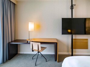 A hotel room with a desk, chair, lamp, TV, and a bed.