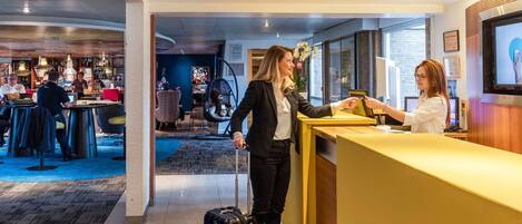 A hotel reception area with a check-in counter, a seating area with tables and chairs, and a bar in the background.