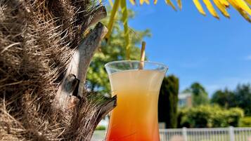 A glass of red and yellow drink next to a pool with a palm tree and a fence in the background.