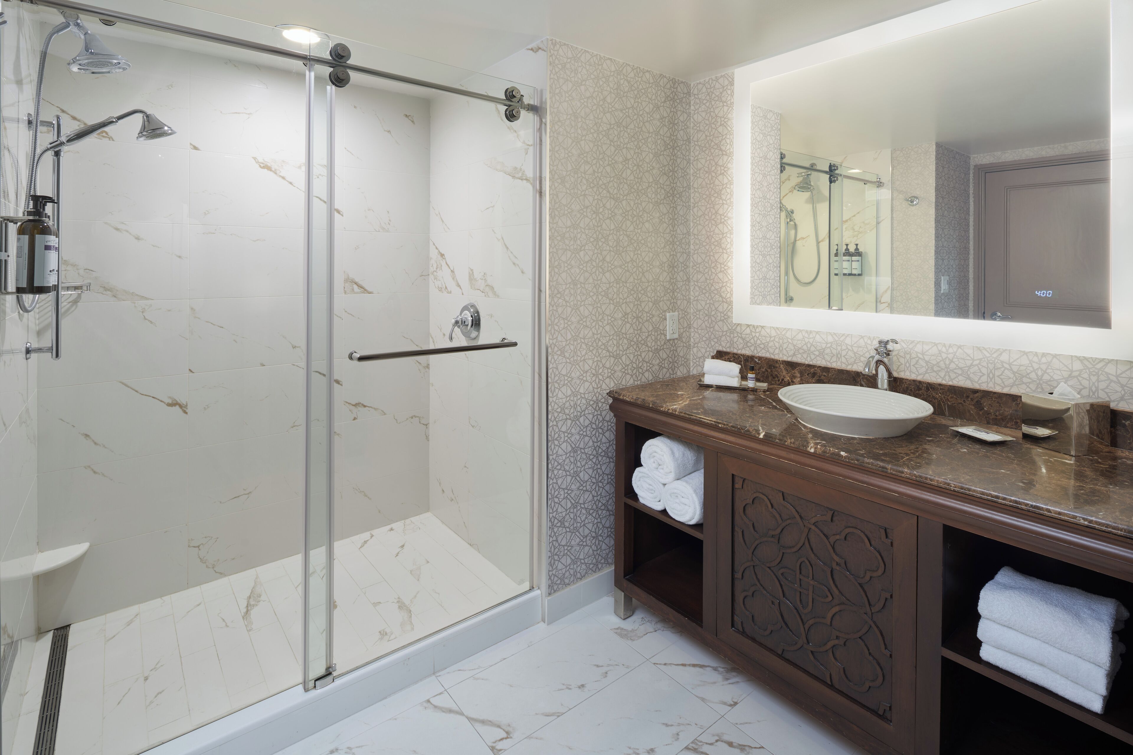 A modern bathroom with a glass-enclosed shower, a marble-tiled floor, a large mirror, and a wooden vanity with a sink and towel storage.