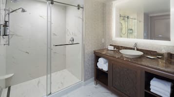 A modern bathroom with a glass-enclosed shower, a marble-tiled floor, a large mirror, and a wooden vanity with a sink and towel storage.