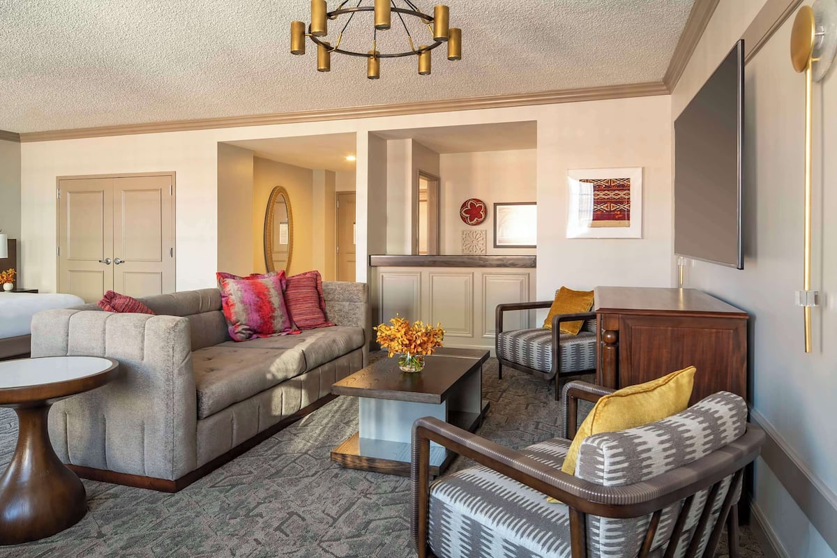 A living room with a sofa, armchairs, a coffee table, and a TV. There is a door leading to another room and a chandelier above.