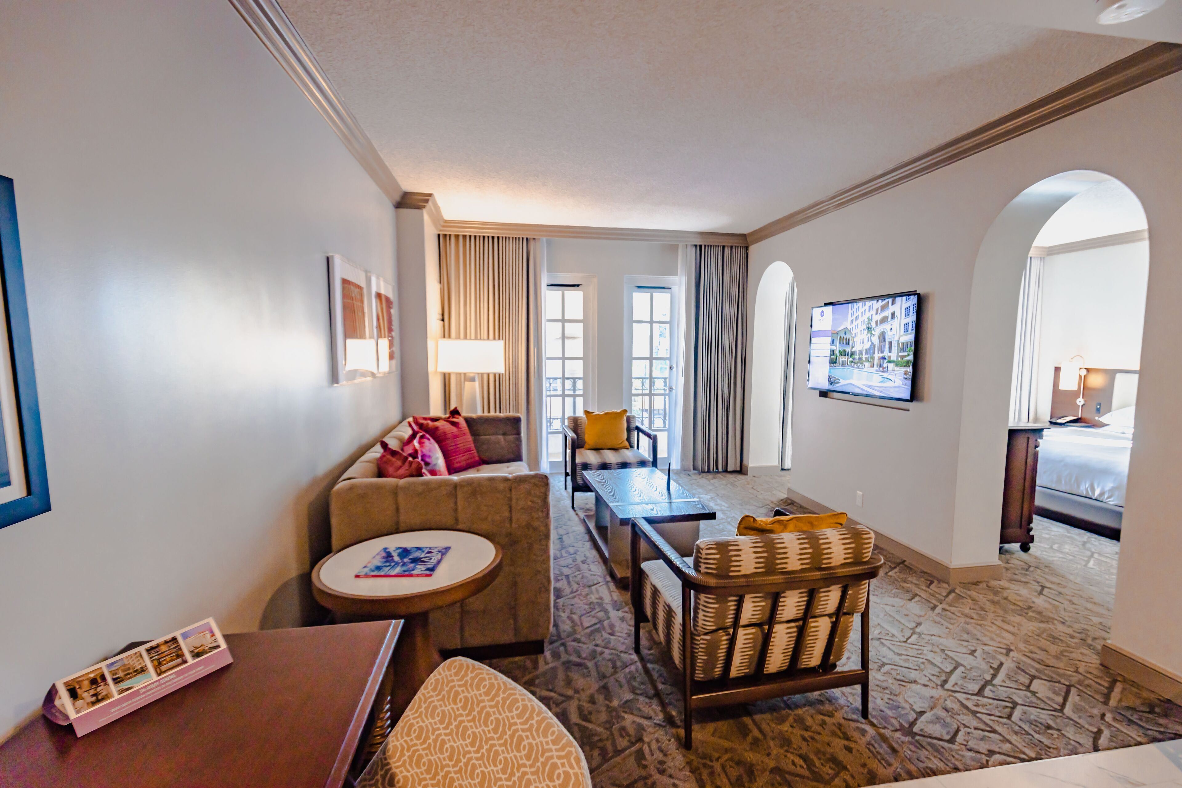 A hotel room with a living area featuring a sofa, a round table, a wicker chair, and a television mounted on the wall.